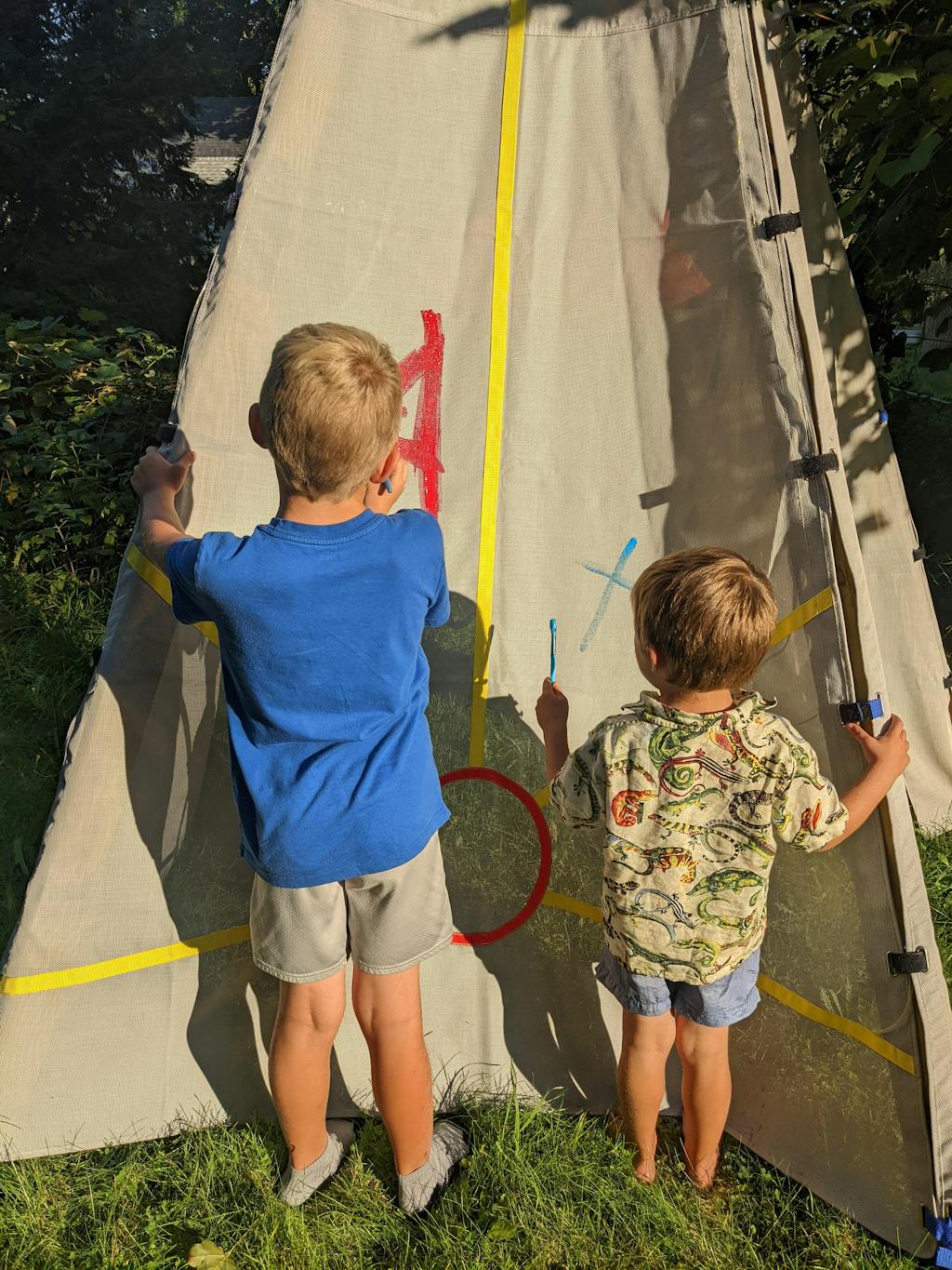 Two children painting a canvas tent outdoors in the sunlight.