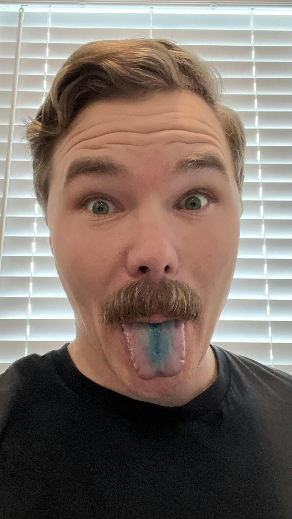 A person with wide eyes sticks out a blue-stained tongue in front of blinds.